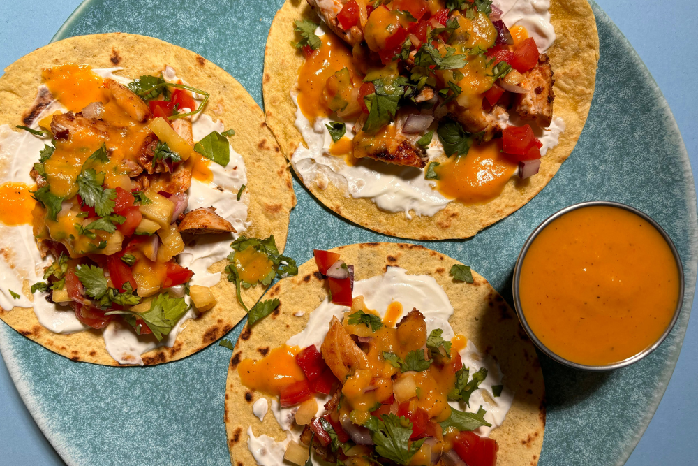  3 tacos on a blue plate, which feature sour cream, chicken, pineapple salsa, and a drizzle of Burnt Pineapple Hot Sauce. There's also a pot of Burnt Pineapple Hot Sauce on the side. 